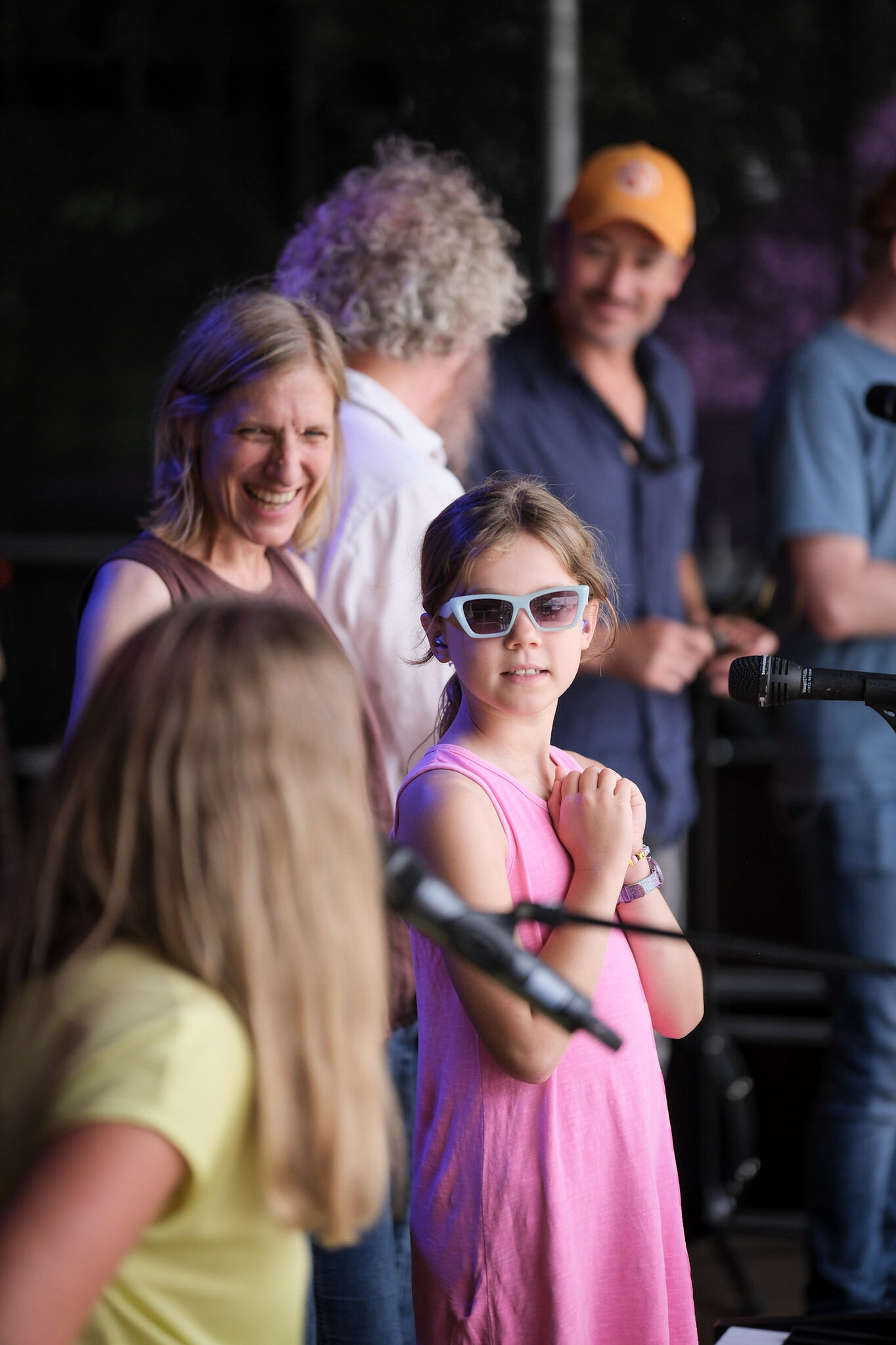 Kinder stehen auf einer Bühne vor Mikrofonen und singen gemeinsam, im Hintergrund Erwachsene.